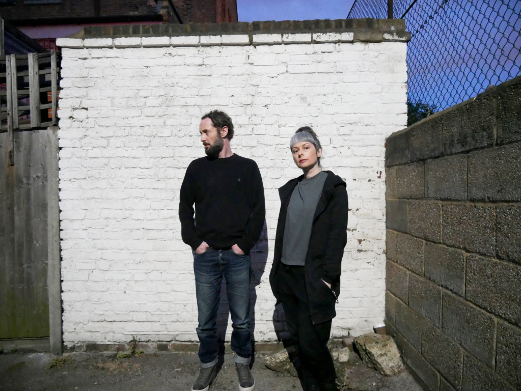 Nicola and Ian of the band 'Stick in the Wheel' stand in front of a brick wall painted white.