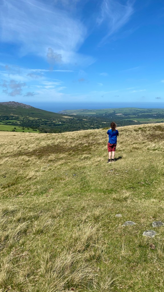 the back of a man with white legs and long dark hair, wearing a blue t-shirt and red shorts standing on short grass. He is looking out to the sea from the top of a hill. There is another mountain in the distance.