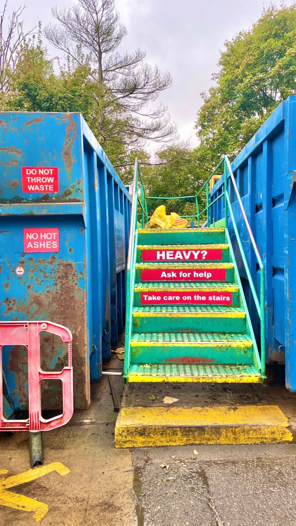 A staircase leads up in between two open-topped shipping containers. The sign reads Heavy? Ask for help. Take care on the stairs.