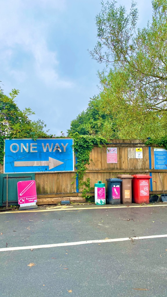 A blue municipal road sign reads One Way, it is next to a fence and some bins on a strip of new tarmac.